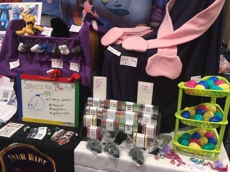 An artist alley table displaying soap, scarves, prints, and squeaky dick keychains