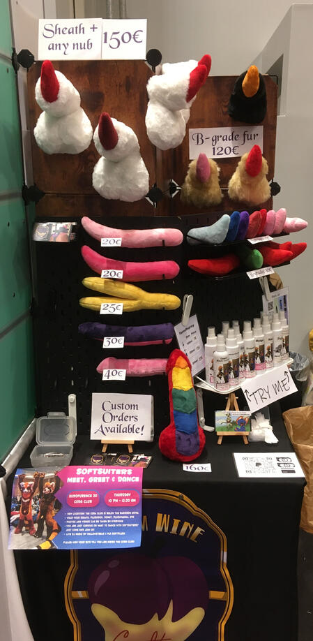 A small dealers den table displaying wearable fursuit sheaths, an array of plush fantasy penises, and fursuit spray